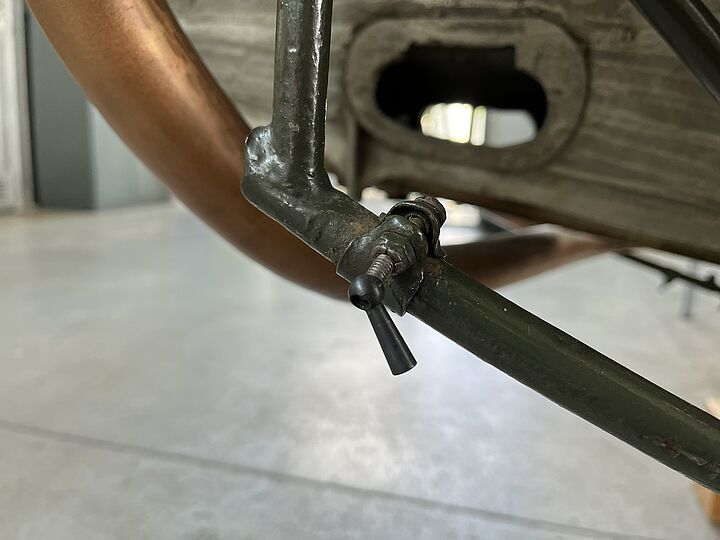 Nahaufnahme eines Metallgestells mit grün lackiertem Rohr, einem angeschraubten kleinen Hebel-/Ventilgriff aus schwarzem Kunststoff und sichtbaren Schrauben sowie einem gebogenen Kupferrohr und Holzteilen im Hintergrund.