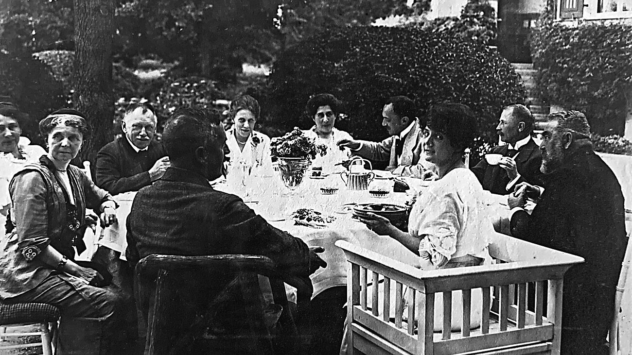 Schwarz-weiß Fotografie einer Teegesellschaft Anfang des 20. Jahrhunderts. Zehn Personen (fünf Männer, fünf Frauen; alle gehobenen Alters) sitzen um einen zum Tee gedeckten, runden Tisch im Garten (Hintergrund Baum, Hecken und Treppen zum Rundbogen-Hauseingang). Blumenbouquet in der Mitte des Tisches. Einige Personen blicken in die Kamera, einige sind in Unterhaltungen.
