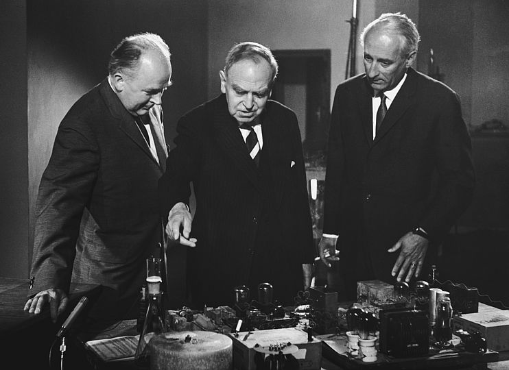 Schwarz-weiß Fotografie. Fritz Straßmann und Otto Hahn erklären Heinz Haber ihre Geräte.