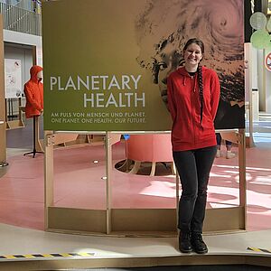 Eine junge Frau mit Zopf und roter Kapuzenjacke steht lächelnd in einer Museumsausstellung vor einer großen Infotafel mit der Aufschrift „PLANETARY HEALTH – Am Puls von Mensch und Planet“.