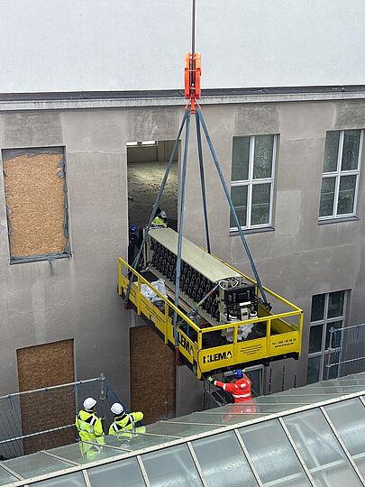 Außen am geöffneten Fenster im Baustellenbereich des Gebäudes hängt die gelbe Transportplattform am Kran und dockt am Fenster an.