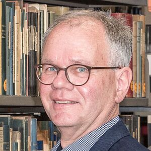 Porträt eines lächelnden älteren Mannes mit kurzen silbergrauen Haaren und braunen runden Brillen, halbseitig zur Kamera gewandt, vor einem Regal mit zahlreichen Büchern.