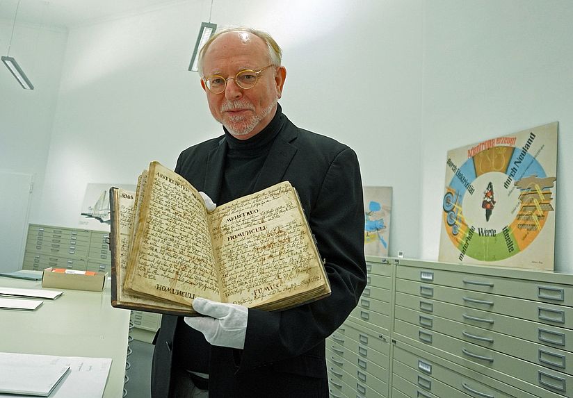 Dr. Wilhelm Füßl hält eine alchemistische Handschrift, dabei trägt er Handschuhe, und zeigt sie der Kamera