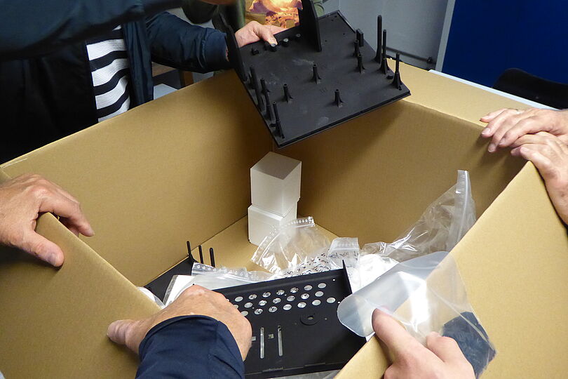 Großer geöffneter Karton auf einem Tisch, mehrere Hände packen darin schwarze Kunststoffteile mit Stiften und gelochte Frontplatte aus, im Karton transparente Plastikbeutel, weiße Schaumstoffpolster und Verpackungsmaterial sichtbar.