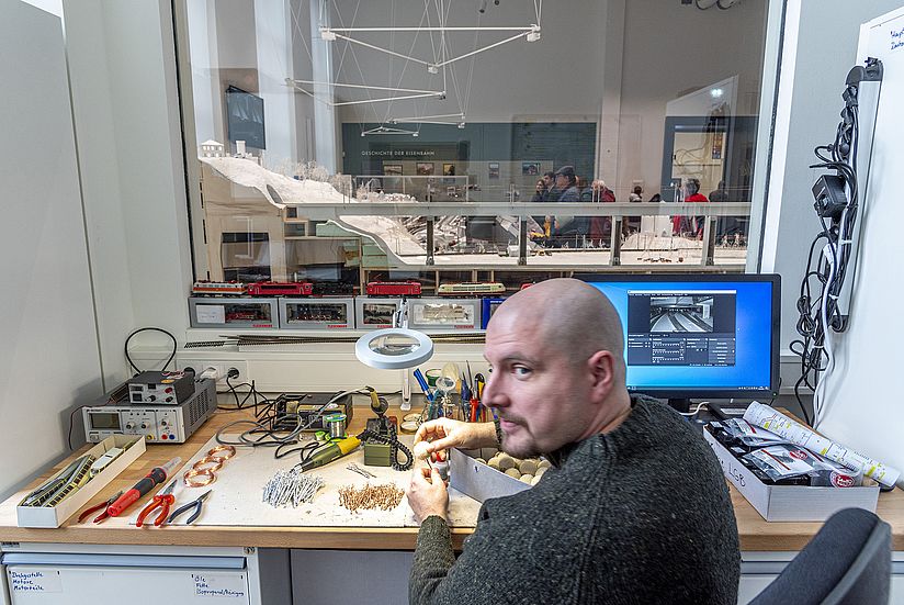 Ein Mitarbeiter sitzt an einem Arbeitstisch, auf dem viele Werkzeuge liegen, und blickt über die Schulter in die Kamera. Er bearbeitet einen Miniaturbaum aus Draht und Schwammgummi. Im Hintergrund durchs Fenster ist die Modellbahnanlage zu sehen.