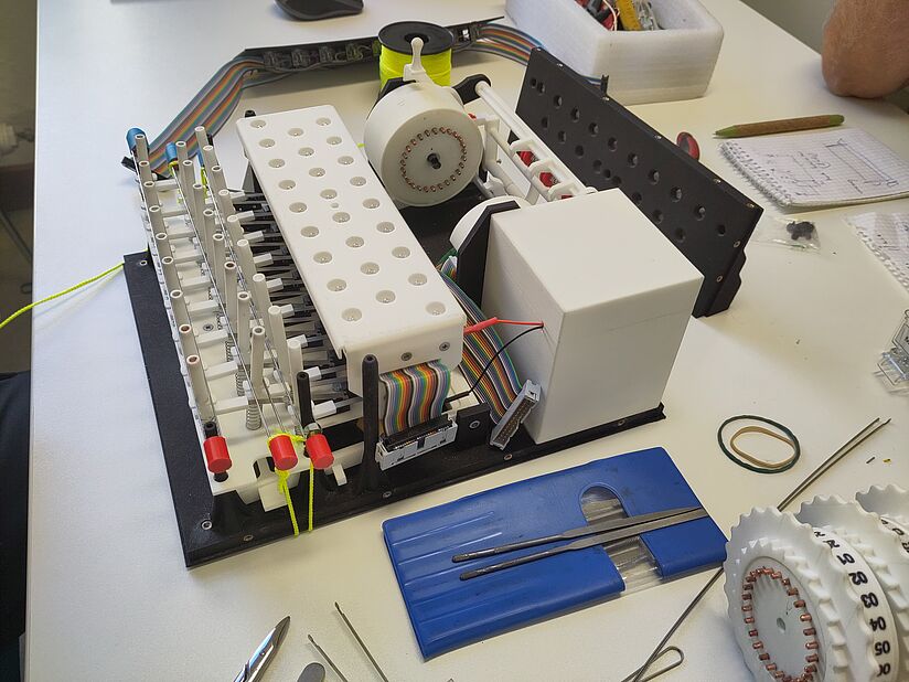 Mechanische Elektronikbaugruppe auf weißer Werkbank: schwarzes Montagebrett mit weißen 3D-gedruckten Bauteilen, Reihen von federnden Stiften und Steckkontakten, mehrfarbiges Flachkabel, großer weißer Würfel als Gehäuse, rundes Zahnkranzrad mit Kupferwicklungen, gelbe Schnur an roten Endkappen, daneben blaue Kunststoffwerkzeugtasche mit Feilen, Notizblock und Gummibänder — Entwicklungs- und Montageumgebung.