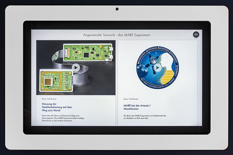 Tablet-Display zeigt eine Webseite zum MARE-Experiment mit links zwei grünen Elektronikplatinen und einem silbernen Flachbandkabel als Bild und Play-Button, rechts ein rundes blau-gelbes Missionslogo (MATROSHKA ASTRORAD RADIATION EXPERIMENT) mit Play-Button; Überschrift „Angewandte Sensorik - das MARE Experiment“ und deutscher Beschreibungstext zu Strahlenmessung und Artemis I.
