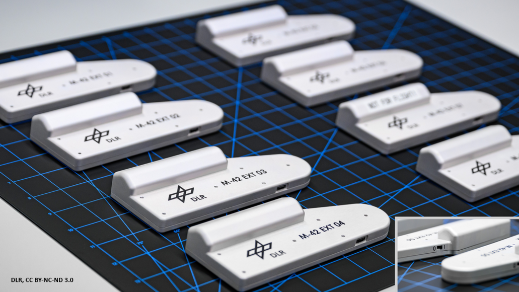 Mehrere weiße, rechteckige Elektronikgehäuse mit abgerundeten Enden und erhöhten Deckeln, beschriftet mit "DLR" und Kennungen wie "M-42 EXT 03/04", angeordnet in Reihen auf einer schwarzen Schneidematte mit blauem Raster; Nahaufnahme zeigt USB-Anschluss und detaillierte Gravuren.