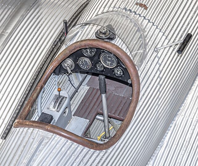Blick von schrägoben in das offene Cockpit eines silbernen alten Flugzeugs. Verschiedene Anzeigeelemente und Hebel sind zu sehen.
