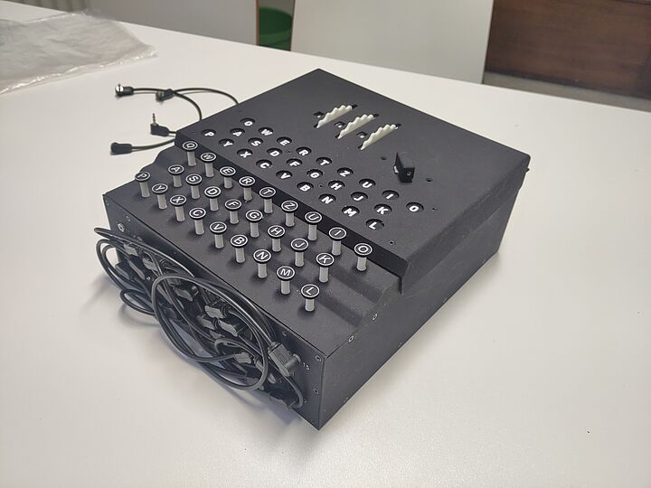 Schwarzes elektromechanisches Gerät mit mehreren Reihen runder, weißen Tasten beschriftet mit Buchstaben (QWERTZ-Layout), drei weißen Schaltern oben, einem Hebel und aufgewickeltem Kabel an der linken Seite, auf weißem Tisch liegend. ENIGMA-Nachbau.
