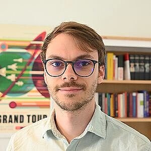 Porträt eines jungen Mannes mit braunen Haaren, leichtem Bart und dunklen Brillenfassungen, vor einem Bücherregal und einem bunten Poster an der Wand.