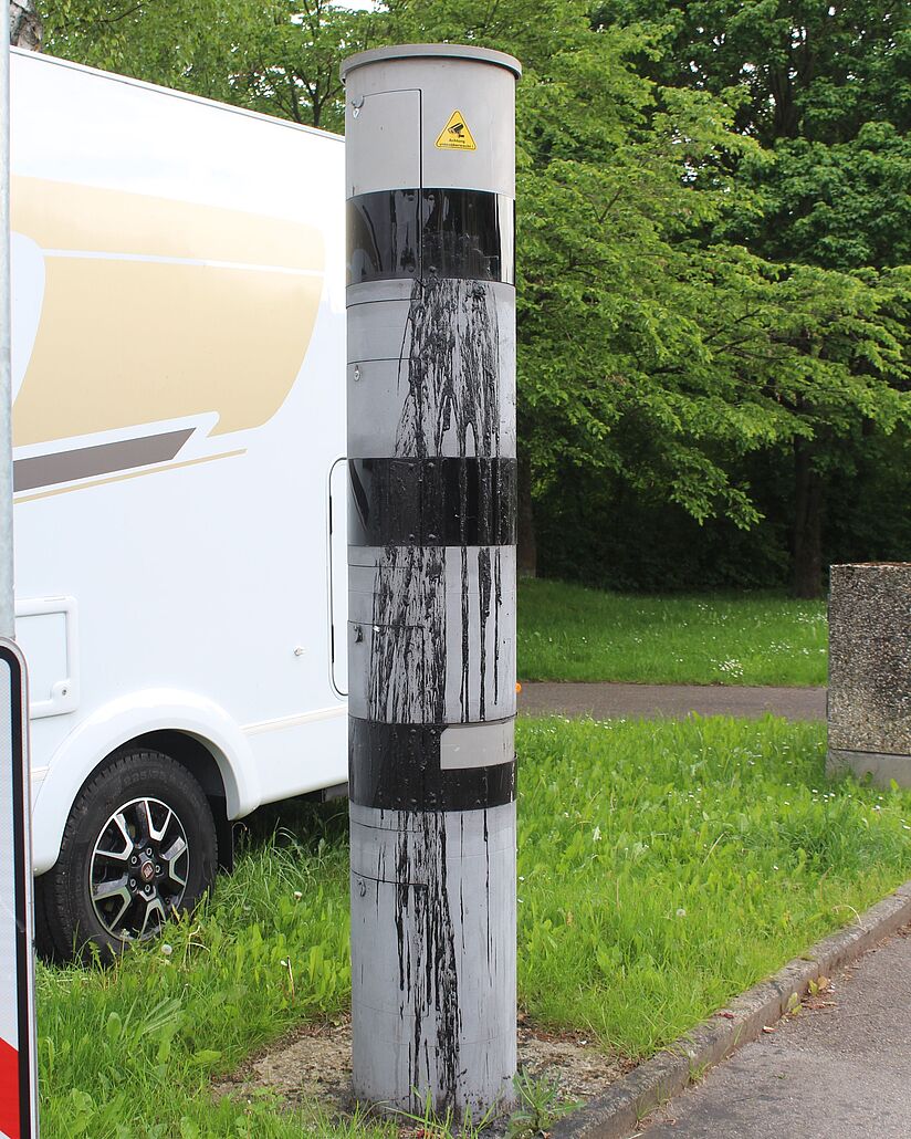 Ca. 1,5 Meter hohe Blitzersäule neben parkendem Van. Man sieht schwarze Farbspritzer und -flächen über die Blitzersäule laufen.