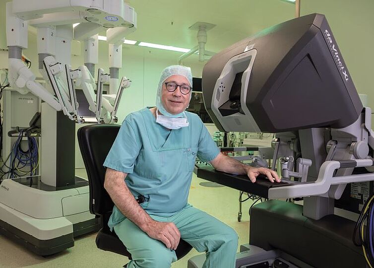 Fotografie eines sitzenden Mannes in OP-Bekleidung. Sitzt vor großem schwarzgrauen Element (Bedienkonsole), im Hintergrund sieht man den vierarmigen OP-Roboter