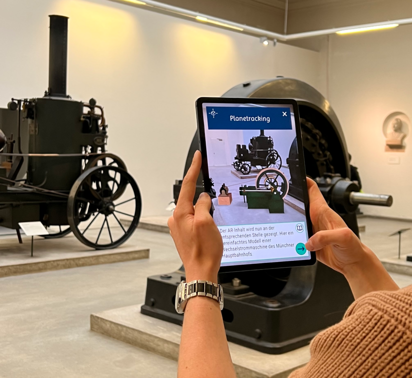 In der Ausstellung mit der AR-App GREIFbAR. Auf einem Tabletscreen ist der Zusatzinhalt "Modell einer Wechselstrommaschine" zu sehen.