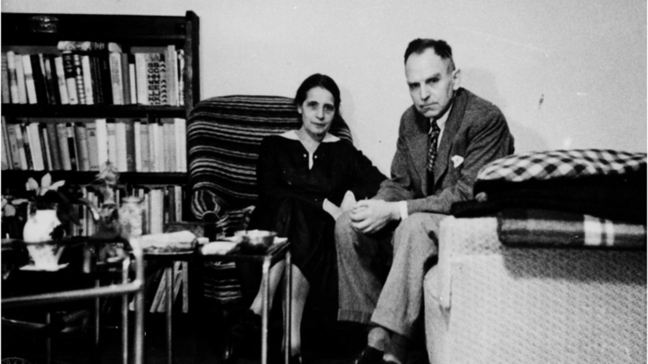 Schwarzweiß Fotografie. Meitner und Hahn sitzend zentral, neben niedrigem Bücherregal und Couch.