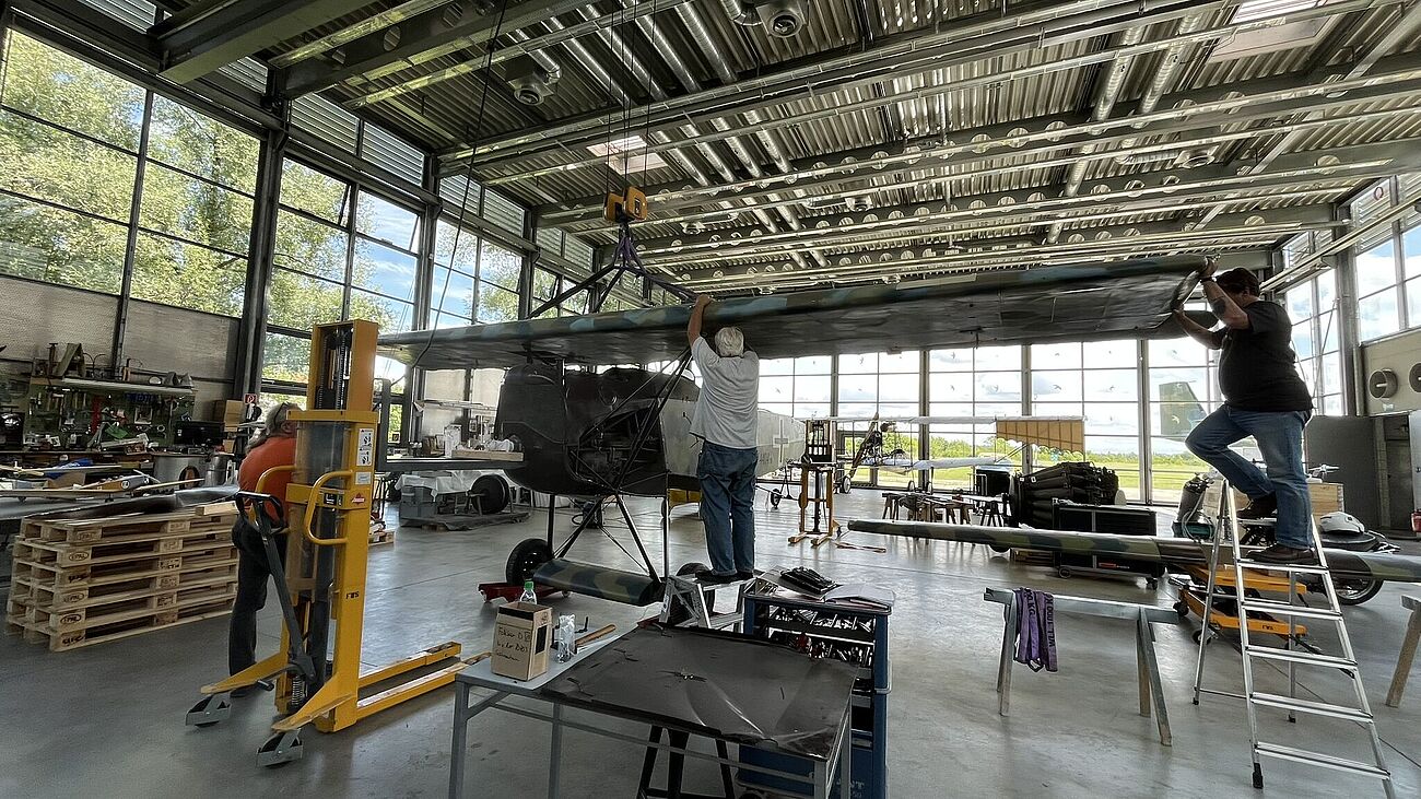 Werkstatthalle mit großem Doppeldecker-Flugzeug in Restaurationsarbeit: zwei Männer stehen an und unter dem oberen Tragflügel, einer auf einer Leiter, zahlreiche Werkzeuge, Hebevorrichtung (gelber Stapler), Paletten, Werkbänke und Ersatzteile verteilt auf Betonboden, große Fensterfront mit Außenblick und sichtbarer Hallendecke mit Metallträgern.