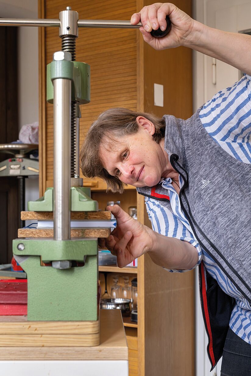 Fotografie einer Frau in einer Werkstatt beim Bedienen eines vertikal angebrachten Schraubstocks. Mit der rechten Hand dreht sie, mit der linken justiert sie das zwischen den Pressbacken angebrachte Buch.