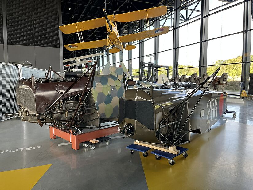 Zwei freistehende Rumpfteile historischer Doppeldeckerflugzeuge in einer Ausstellungshalle: links ein brauner Motor- und Cockpitrahmen mit freiliegendem Motor auf einem orangen Rollenpodest, rechts ein olivgrüner Rumpf mit der weißen Nummer "266" und rotem Kokardenmarkierung auf einem blauen Wagen; im Hintergrund hängender gelber Doppeldecker, große Fensterfront und weitere Exponate.