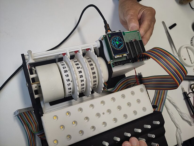 Nahaufnahme eines teilzerlegten elektronischen Geräts mit weißen 3D-gedruckten Kunststoffteilen, vier beschrifteten Ziffern-Rollen (mit Zahlen 00–29), einer kleinen gesteckten Leiterplatte mit OLED-Display, bunten Flachbandkabeln, einer LED-Matrixplatte mit 20 einzelnen LED-Linsen und mehreren weißen Druckknopfstangen; Hand hält die Leiterplatte, Hintergrund helle Werkbank mit Werkzeugen.