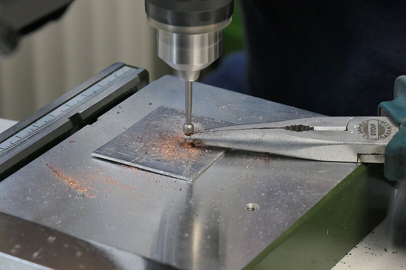Nahaufnahme einer Metallfräs- oder Bohrvorrichtung: ein rotierender Fräser bohrt ein kleines Loch in eine quadratische Metallplatte, feine kupferfarbene Späne liegen um die Bohrstelle, rechts greifen lange Flachzangen die Platte, im Hintergrund Teil einer Messskala und einer Metallarbeitsfläche sichtbar.