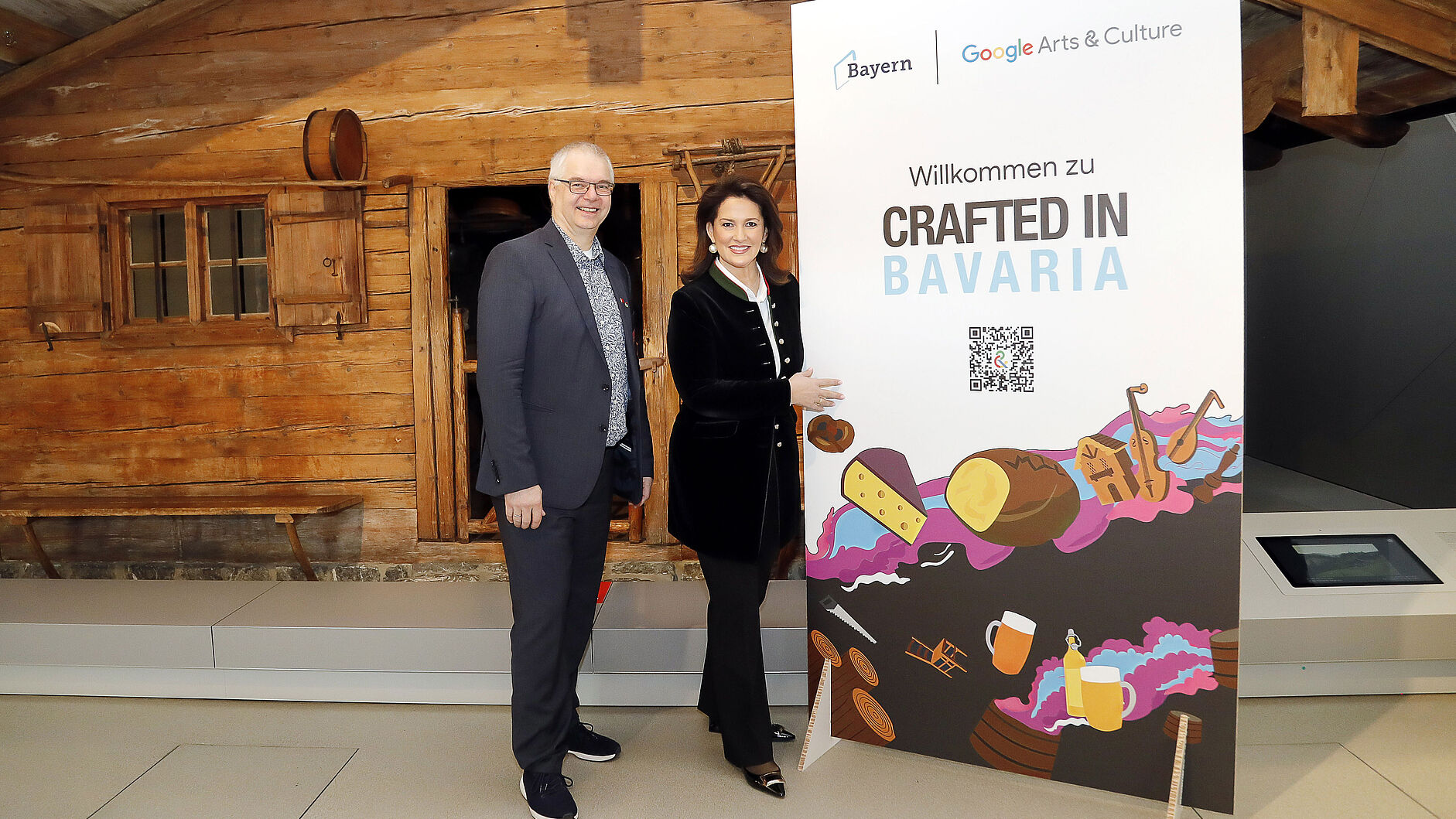 Wieland Holfelder und Michaela Kaniber stehen vor der Almhütte und zeigen auf ein Plakat mit der Aufschrift Crafted in Bavaria.