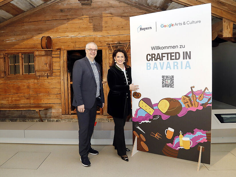 Wieland Holfelder und Michaela Kaniber stehen vor der Almhütte und zeigen auf ein Plakat mit der Aufschrift Crafted in Bavaria.