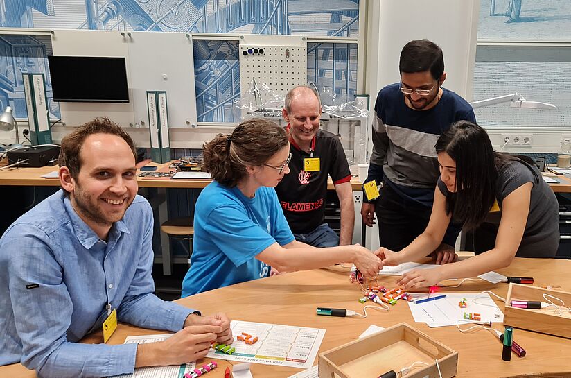 Fünf Personen arbeiten Steckverbindungen an einem Tisch. Vier sind in der Gruppe beim Tüfteln, ein Mann blickt in die Kamera.