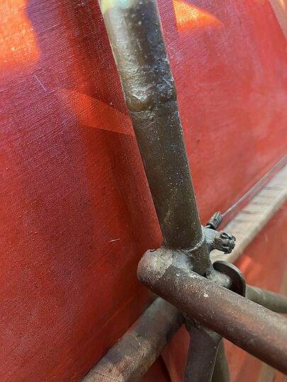 Nahaufnahme eines rostigen Rahmens aus Metall vor rotem Hintergrund, mit sichtbaren Lötstellen und Klemmschraube, schmutzig und korrodiert.