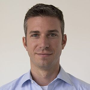 Ein Mann mit verschwommenem Gesicht vor hellem Hintergrund, trägt hellblaues Hemd über weißem T‑Shirt, nur Kopf, Hals und Schultern sichtbar.