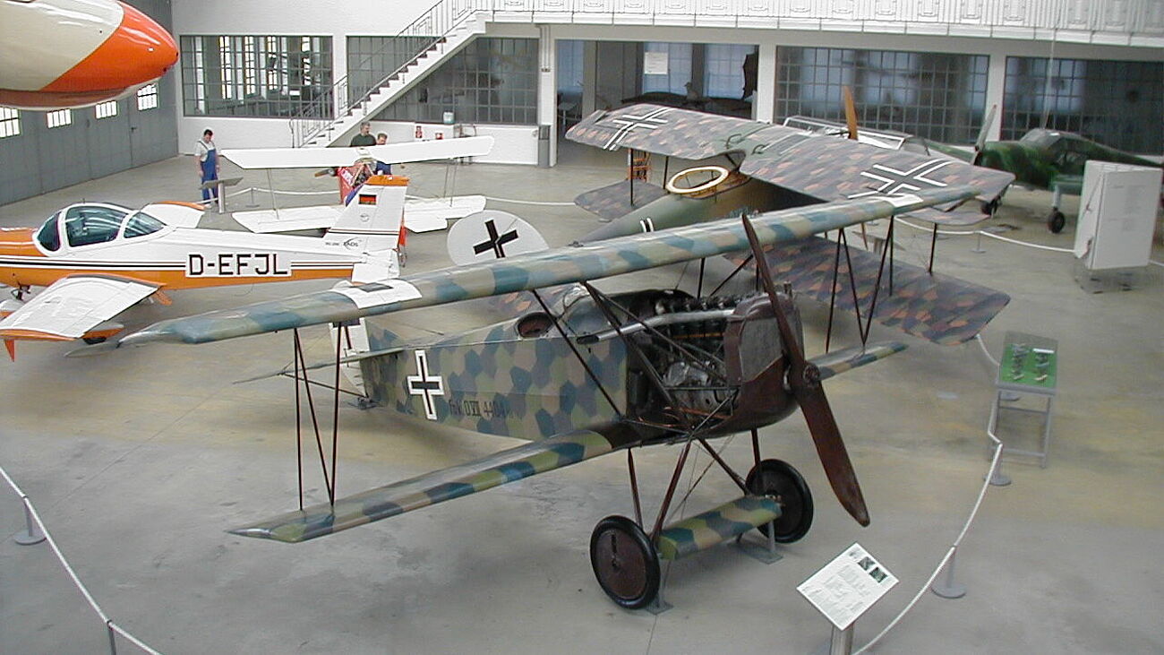 Mehrere historische Flugzeuge in einer hellen Ausstellungshalle: im Vordergrund ein deutsch-kamouflierter Doppeldecker mit Holzpropeller und schwarzem Balkenkreuz, dahinter ein weiterer bemalter Doppeldecker und ein weiß-oranges Sportflugzeug mit Kennzeichen D-EFJL, umgeben von Absperrseilen und Informationsschildern.
