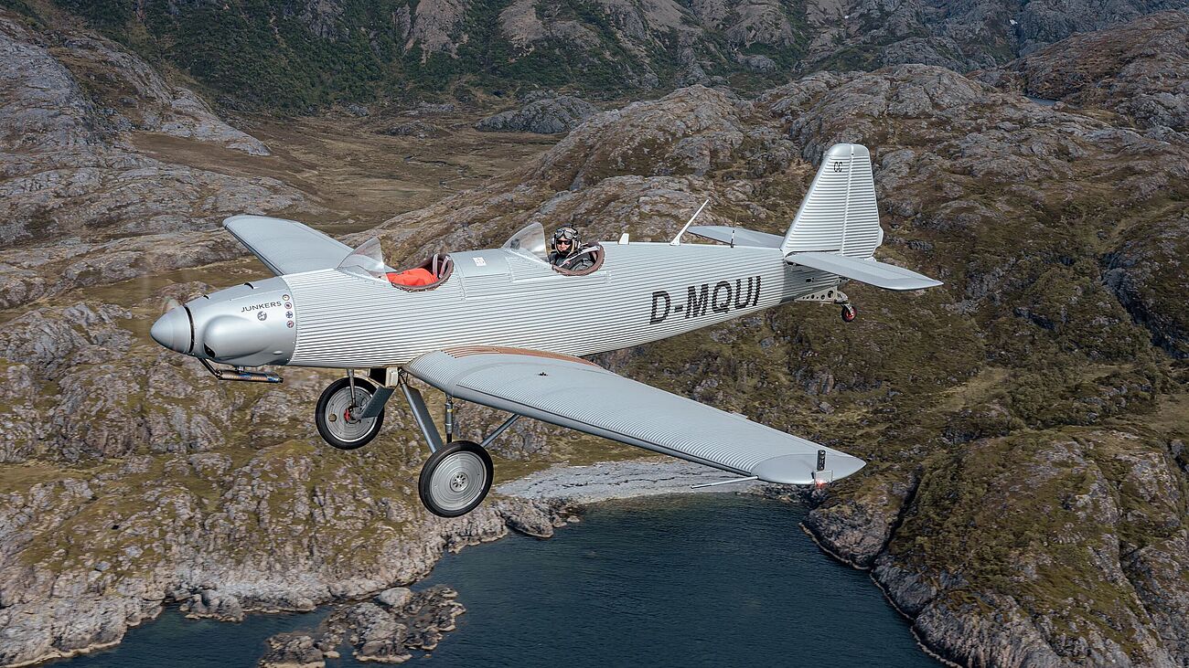 Silbernes, metallenes Flugzeug über bergiger Landschaft mit Wasser im unteren Bildteil. Zwei Sitze hintereinander mit offenem Verdeck. Pilot sitzt in hinterem Sitz.