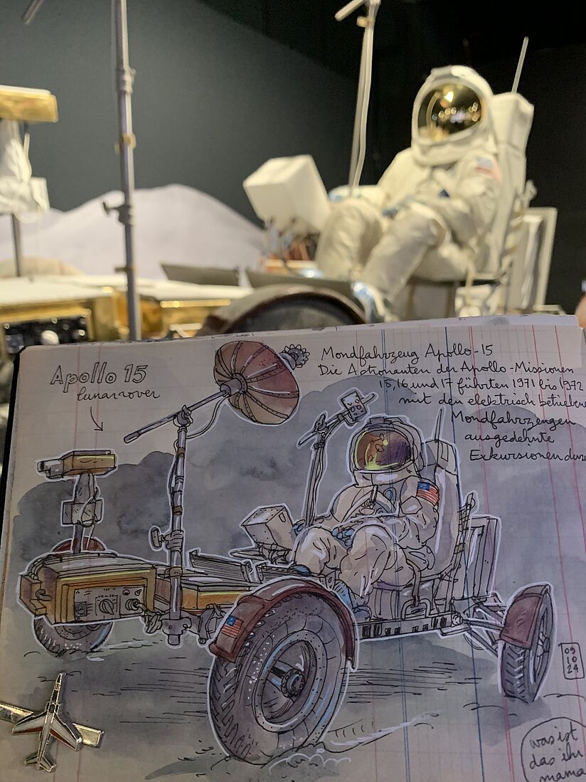 Ein gezeichntetes Bild des Mondautos wird vor das ausgestellte Mondauto gehalten. 