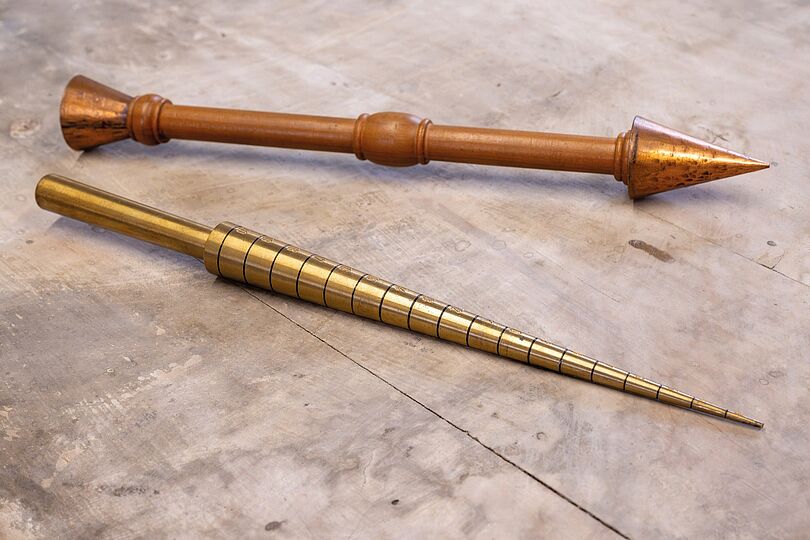 Fotografie von zwei länglichen Instrumenten. Das obere ist aus Holz und ist pfeilförmig mit einer kleinen Ausbuchtung in der Mitte. Das untere sieht aus wie das vordere Ende einer Lanze und ist metallen gelblich mit schwarzen Kreisen um die Spitze.