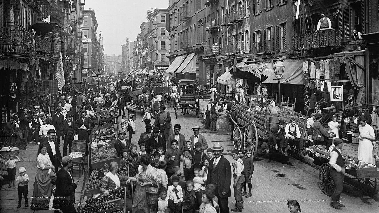 Schwarzweiß Fotografie einer Straße in einer Großstadt. Viele Menschen, Händler, Kinder, eine Gruppe schaut in die Kamera.