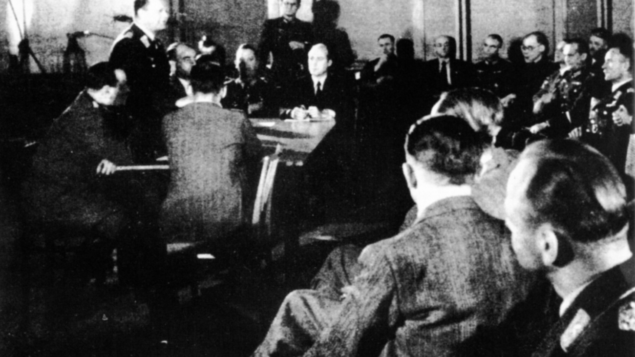 Schwarzweiß Fotografie. Gruppe von Männer, meist in Uniform und sitzend. In der Mitte des Raums ein Tisch, darum wiederum sechs Männer.