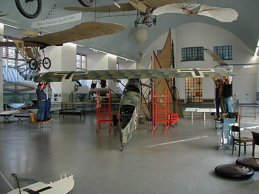 Im Inneren eines Luftfahrtmuseums: ein großes, in Tarnfarben lackiertes historisches Flugzeug von vorne mit ausgebreitetem Oberflügel, darunter rote Gerüste und Werkbühnen, zwei Besucher stehen auf Podesten und berühren die Tragfläche, weitere frühe Fluggeräte und Modellsegel im Ausstellungsraum sowie Museumseinrichtungen und Stühle im Vordergrund.
