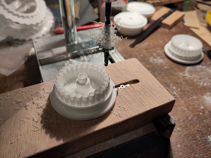 Werkstattaufnahme eines Bohrers über einer weißen, runden Kunststoff- oder 3D-gedruckten Zahnradscheibe mit zahnförmigen Rippen, befestigt auf einem Holzbrett; feine weiße Späne hängen am Bohrer und liegen auf Werkbank und Bauteil, im Hintergrund weitere ähnliche Kunststoffteile und Werkzeuge.