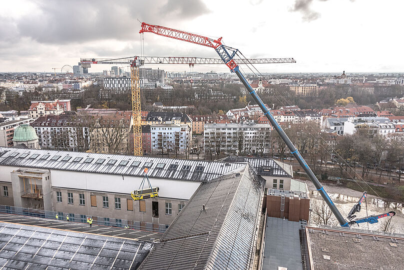 Der Gezeitenrechner hängt an einem Kran auf einer Plattform nahe einem Fenster, während Personen auf einem gegenüberliegenden Dach den Kranführer per Funk einweisen.