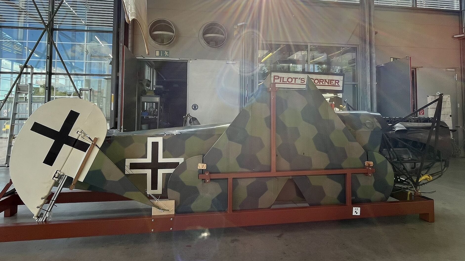 Rumpfabschnitt eines historischen Flugzeugs mit sechseckiger Tarnlackierung in Grün-, Braun- und Olivtönen auf einem roten Transportgestell, sichtbares Heck mit schwarzem Balkenkreuz auf weißem Kreis, im Hintergrund große Hallenfenster mit Sonnenlicht und Schild „Pilot’s Corner“.