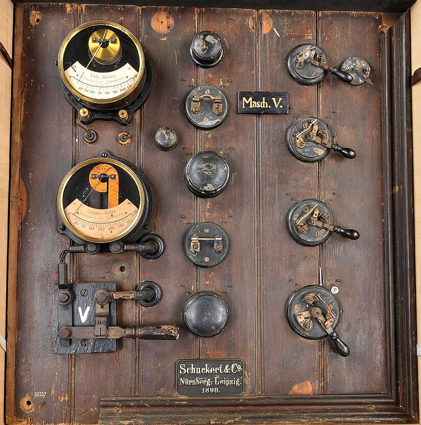 Schalttafel frontal fotografiert. Links oben Volt- und Amperemter in Messingverkleidung. Diverse Bedienelemente auf dunklem Holz.