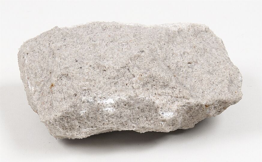 Graubeiger Sandsteinbrocken mit feinkörniger, leicht rauer Oberfläche. Das Gestein zeigt eine kompakte Struktur und besteht aus fest verbackenen Sandkörnern.