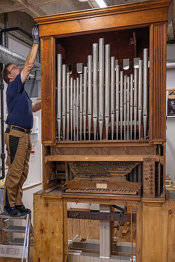 Fotografie von einem Mann, wie er neben einer freistehenden kleinen Orgel auf einer Staffelei steht. Mit der Hand greift an den oberen Teil des Rahmens der Orgel.