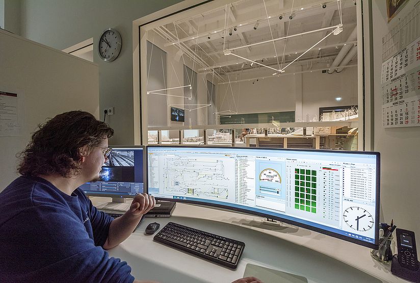 Ein Mann bedient einen PC. Auf dem Monitor ist eine komplexe Softwareoberfläche mit einem schematischen Gleisplan der Modellbahn zu sehen. Im Hintergrund sieht man durchs Fenster die Modellbahnanlage.