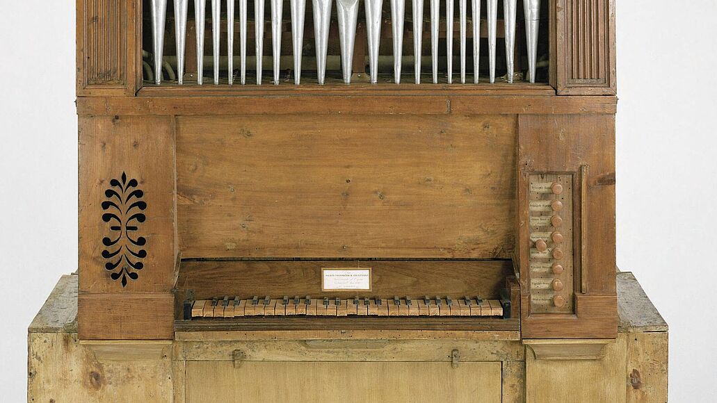 Frontale Fotografie einer kleinen, alleinstehenden Kirchenorgel. Man sieht die silbernen Pfeifen in einem aufgesetzten Kasten, die hölzerne Klaviatur (4 Oktaven), die Register seitlich daneben und die Fußpedale (eine Oktave).