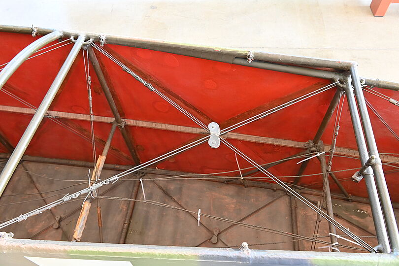 Aufsicht auf das Metallgestell einer Plane: roter, leicht verschmutzter Bespannungsstoff, Stahlrohre und Querstreben mit verspannten Stahlseilen, Umlenkrolle in der Bildmitte und Klemmen/Schlaufen zur Befestigung.
