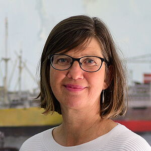 Eine Frau mit verschwommenem Gesicht steht vor einem großen maritimen Wandbild mit Schiffen und Kränen und hält zusammengefaltete Pläne oder ein gerolltes Papier; sie trägt einen weißen, strukturierten Pullover und graue Hosen.