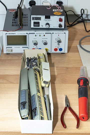 Auf einem Arbeitstisch steht ein kleiner Karton, in dem sich zerlegte Modelle von Miniaturstraßenbahnen befinden. Ringsherum liegen Werkzeuge.