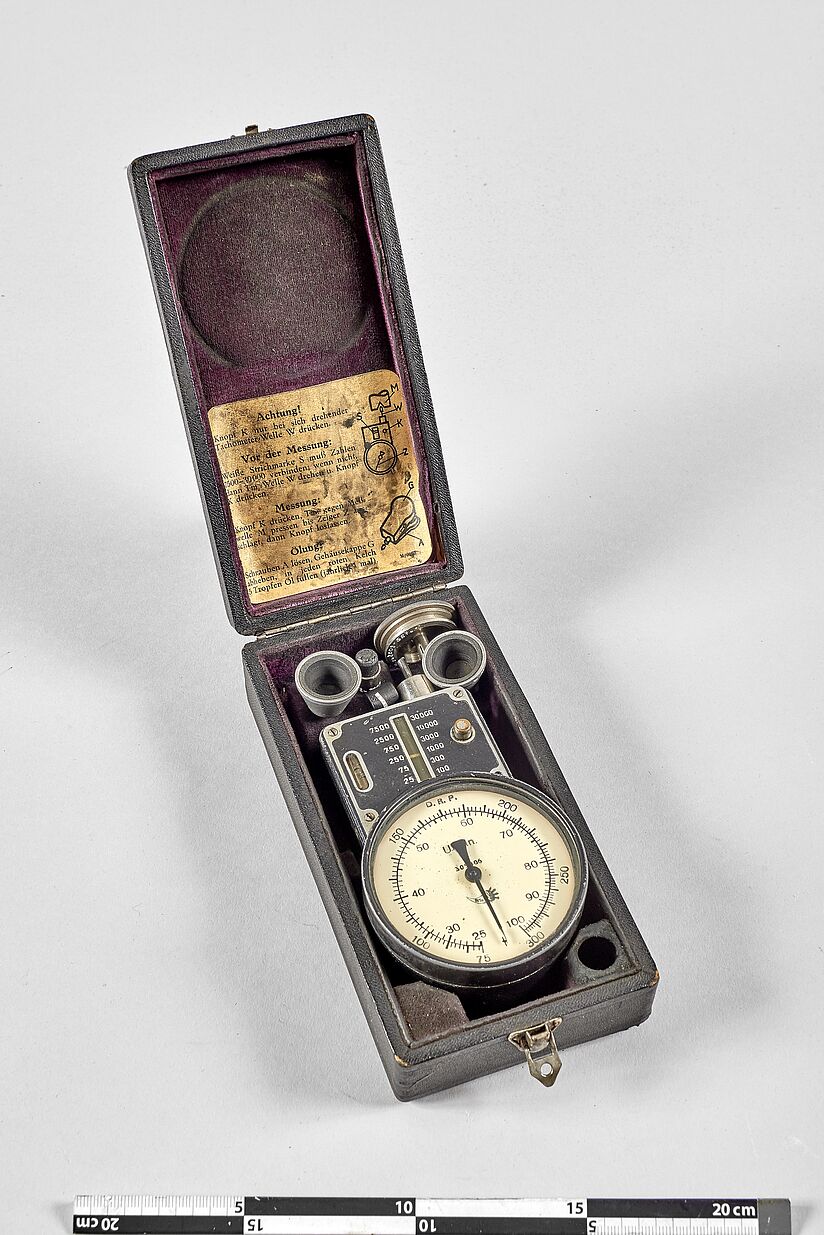 Geöffnetes Etui. Oben ein bedruckter Zettel eingelegt, unten eingelegt: rundes Anzeigefeld mit Zeiger und verschiedenes technisches Zubehör.