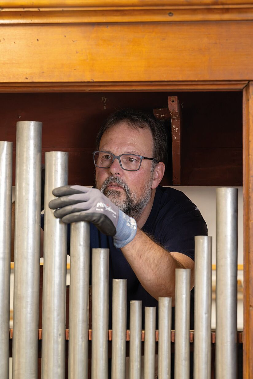 Fotografie. Ein Man blickt konzentriert auf eine Orgelpfeife und hält diese mit einem Handschuh fest.
