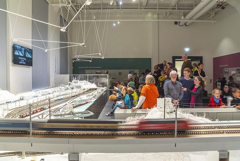 Im Vordergrund fährt ein Modellbahnzug so schnell vorbei, dass er wegen seiner Geschwindigkeit auf dem Bild verschwommen erscheint. Dahinter öffnet sich der Blick über die Modellbahnanlage, die vom Publikum besichtigt wird